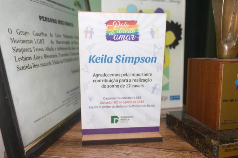 Foto de um troféu. No topo dele, está escrito “Pelo direito de amar” sobre uma bandeira arco-íris. No centro: "Keila Simpson - Agradecemos pela importante contribuição para a realização do sonho de 12 casais. Casamento coletivo LGBT - Salvador 02 de agosto de 2019. Escola Superior da Defensoria Pública da Bahia". No rodapé, encontra-se a logo da Defensoria Pública da Bahia.