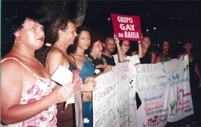 Digitalização de foto analógica da manifestação contra assassinato de pessoas LGBTs. Na foto, é possível ver várias pessoas alinhadas a partir da diagonal inferior esquerda, segurando faixas de protesto.