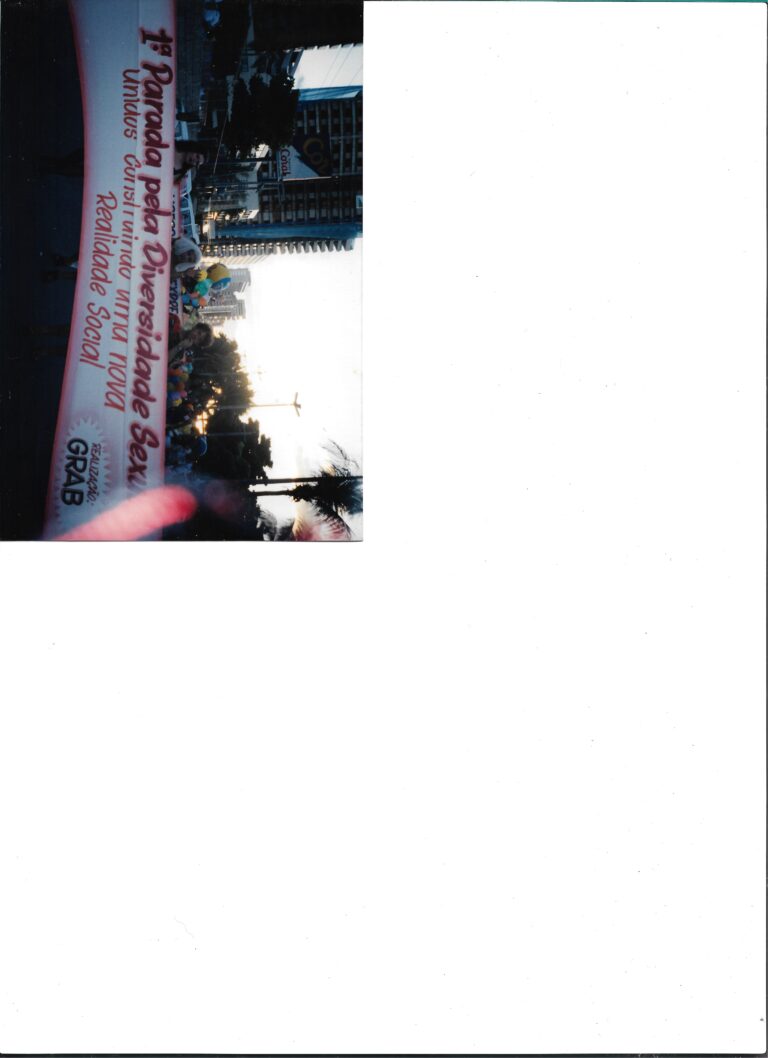 Digitalização de foto analógica do encerramento do VII ENTLAIDS. A foto ocupa o canto inferior esquerdo da página A4, deixando o espaço restante em branco. Não possui marcas. Na foto, é possível ver pessoas carregando uma faixa com os dizeres “1a parada pela Diversidade Sexual”.