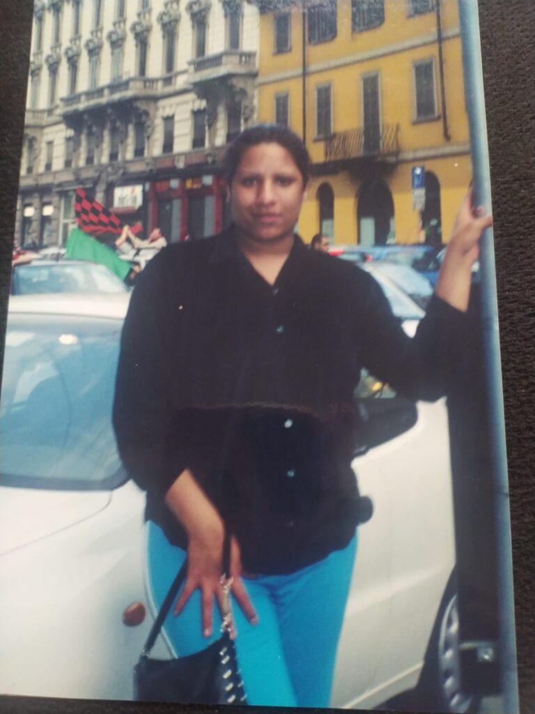 Foto analógica digitalizada, um pouco torta. É possível ver uma mesa ou tampo escuro atrás da foto analógica. Não há marcas visíveis na fotografia. Kelly Passos está no centro da foto apoiando braço esquerdo em um poste, vestindo calça azul e camisa preta de frio de botões, segurando uma bolsa preta, na rua de uma cidade durante o que parece ser uma carreata ou comemoração com diversos automóveis.