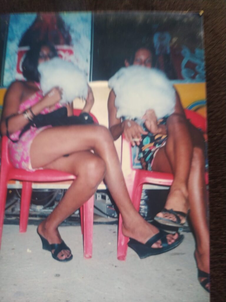 Digitalização de uma foto analógica, em qualidade não muito boa. A foto está deslocada no quadro e borrada. Nela, Kelly Passos (esquerda) e Açucena (em memória) (direita) vestindo vestidos florais coloridos e sentadas em cadeiras de plástico vermelhas, ambas comendo algodão doce branco que cobre seus rostos.