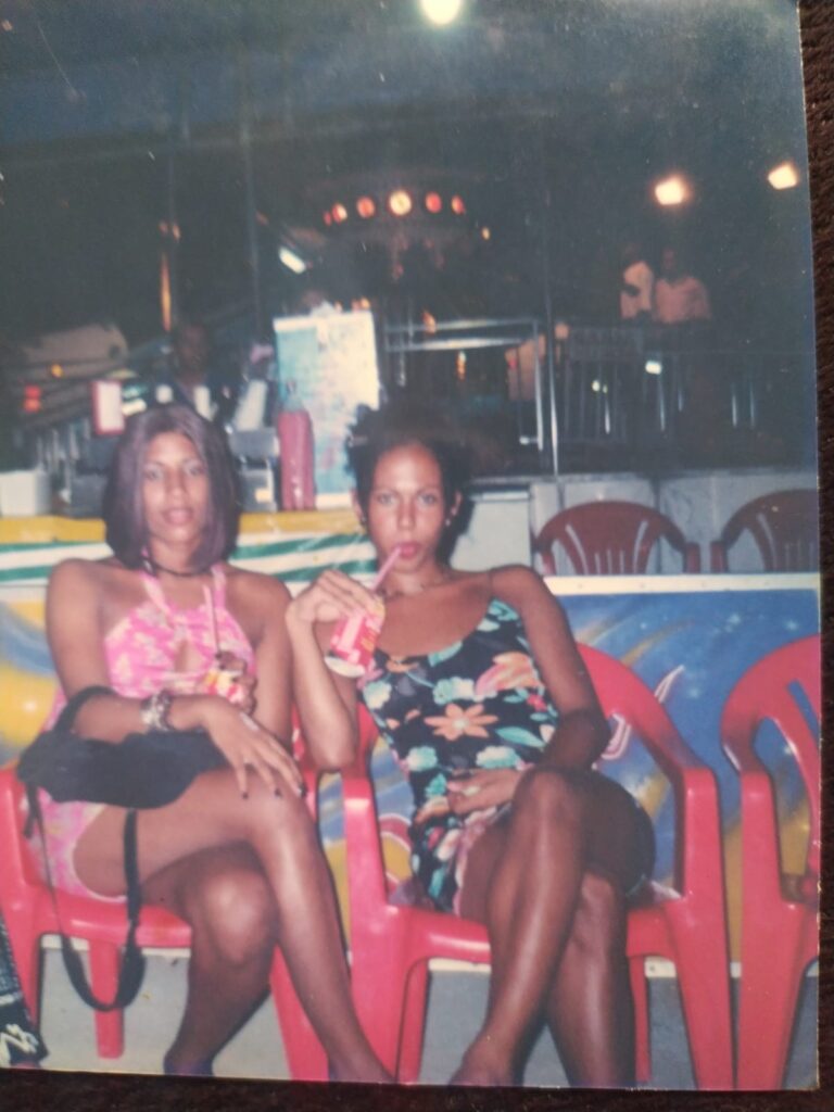 Foto analógica digitalizada, um pouco deslocada. É possível ver o tampo da mesa à direita e abaixo. Não há marcas visíveis na foto. Nela, estão Kelly Passos (esquerda) e Açucena (em memória) (direita) vestidas em vestidos de flores e sentadas em cadeiras de plástico vermelhas, em um parque de diversões. Uma das pessoas bebe uma coca cola de canudo.