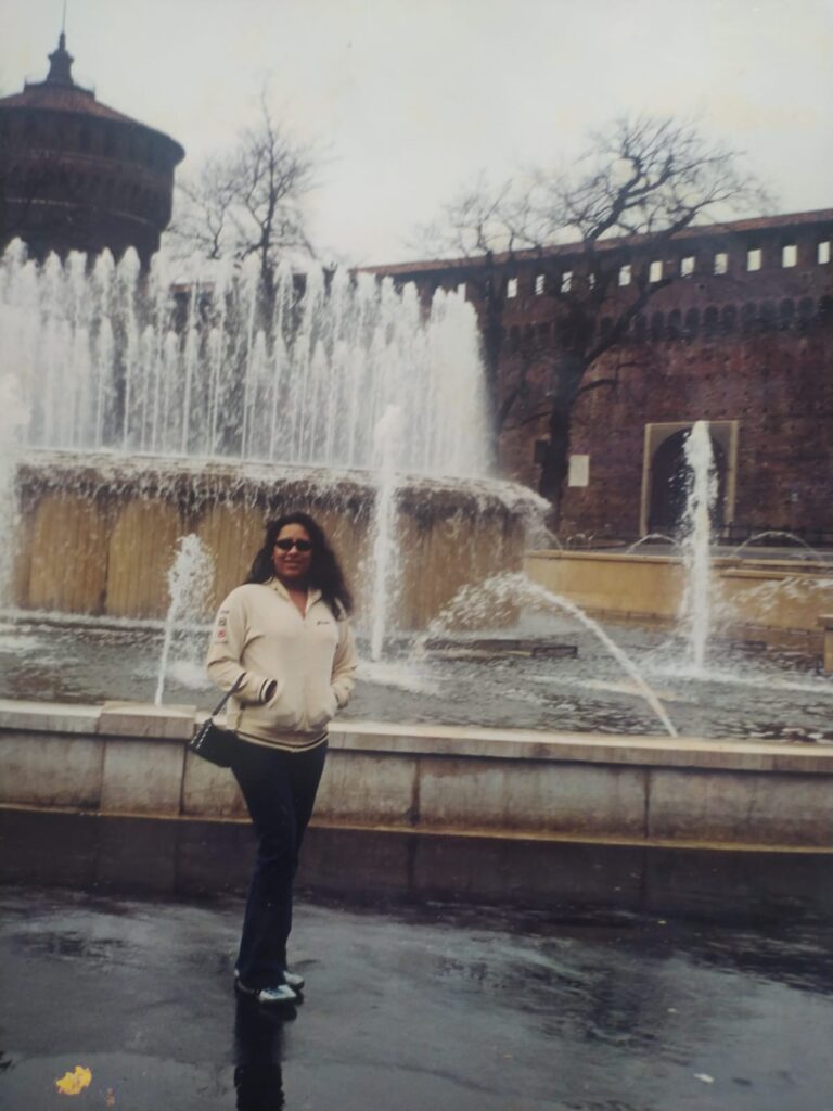 Digitalização de foto analógica. Kelly Passos está de pé, com casaco branco/creme, óculos escuros e jeans, em frente a um chafariz. Não há marcas visíveis na foto.