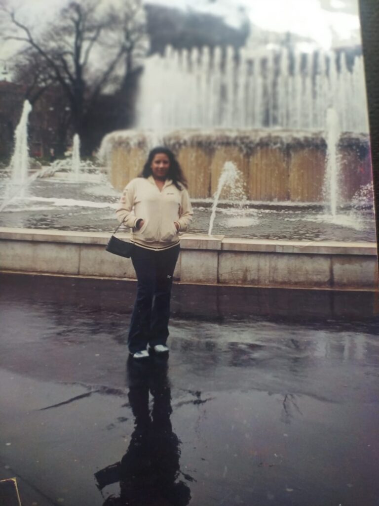 Foto analógica digitalizada, um pouco deslocada no quadro. É possível ver, no canto superior direito, um pedaço do tampo de mesa. Existe uma marca na foto, no canto inferior esquerdo. No centro do quadro, está Kelly Passos de casaco branco ou creme e segurando pequena bolsa preta no braço direito, de pé em frente a um chafariz.