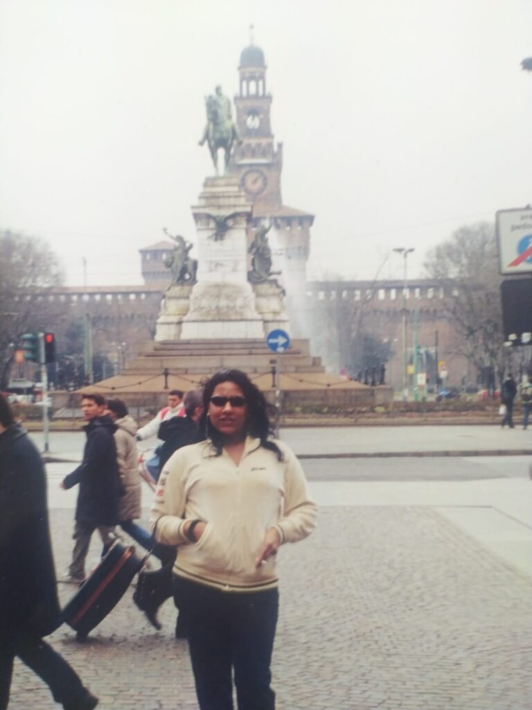 Foto analógica digitalizada. Não há marcas visíveis na foto. Kelly Passos aparece dos joelhos para cima, de casaco branco/creme e óculos escuros está de pé. Atrás dela há uma praça com uma estátua de homem cavalgando.