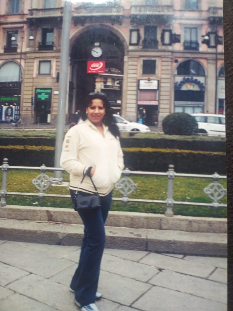 Foto analógica digitalizada, um pouco deslocada no quadro. É possível ver o tampo da mesa no canto direito da foto. No centro, está Kelly Passos de casaco branco/creme e calça jeans, segurando uma bolsa preta. Atrás dela há um prédio que parece comercial.