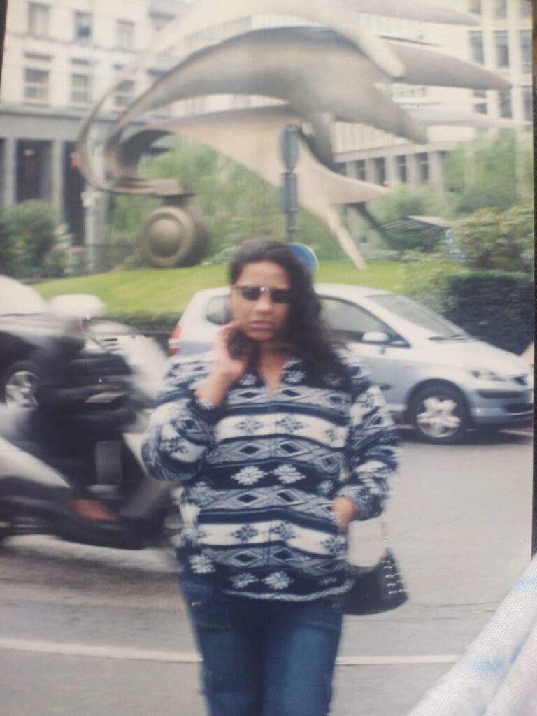 Foto analógica digitalizada. A foto está um pouco precária, borrada. Nela, Kelly Passos está no centro usando óculos escuros e um casaco estampado azul e branco/cinza. É possível ver uma rua ou um estacionamento de carros atrás dela e ao fundo uma escultura metálica com formas alongadas que parecem dedos de uma mão.