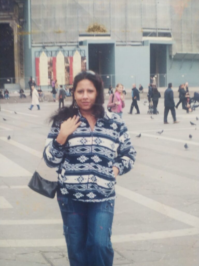 Foto analógica digitalizada. Não há marcas visíveis na foto. No centro dela, Kelly Passos veste bolsa e casaco estampado azul e cinza/branco. Ela está no pátio de um prédio com arquitetura antiga. Há pombos e pessoas no pátio, à distância.