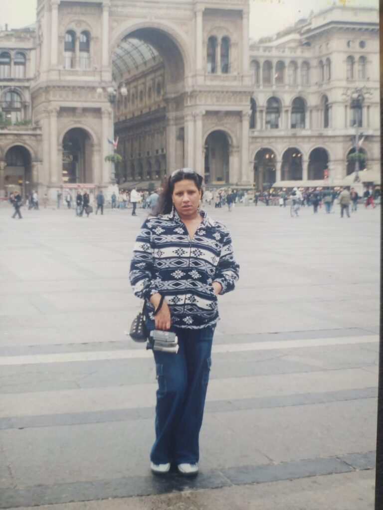 Foto analógica digitalizada, um pouco deslocada. É possível ver um pedaço de mesa no canto inferior direito. Não há marcas visíveis na foto. No centro dela, Kelly Passos veste casaco estampado com cores azul e cinza/branco, na frente do pátio de um prédio histórico.