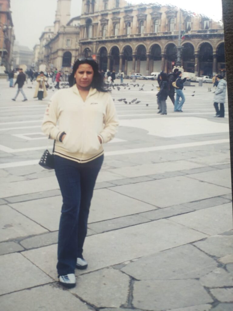 Foto analógica digitalizada, um pouco deslocada no quadro. É possível ver, no canto direito, um pedaço do tampo da mesa. No centro da foto está Kelly Passos vestindo um casaco bege e calça jeans, segurando pequena bolsa no braço direito. Atrás dela está o Coliseu ou um prédio histórico com arquitetura semelhante.