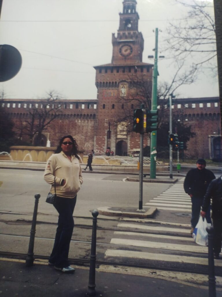 Foto analógica digitalizada. O quadro da foto está um pouco deslocado, e é possível ver um pedaço do tampo da mesa. Na foto, há Kelly Passos vestindo casaco branco/creme e óculos escuros. Ela está na frente de uma praça com construção similar a castelo com um relógio.