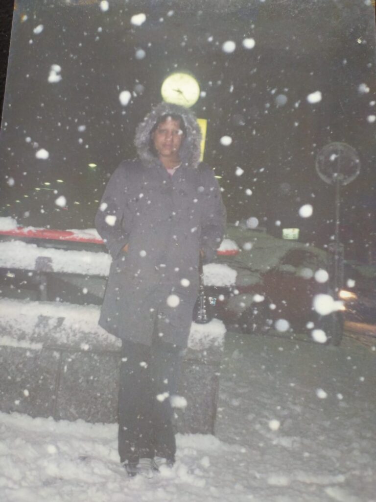 Foto analógica digitalizada, um pouco fora de quadro. A foto está um pouco dobrada no canto superior esquerdo. Não há outras marcas visíveis. No centro da foto, há Kelly Passos veste casaco corta vento e capuz e está com as mãos dentro dos bolsos. É possível ver muita neve no chão, no carro logo atrás, no ar ao redor dela e na frente da lente da câmera.