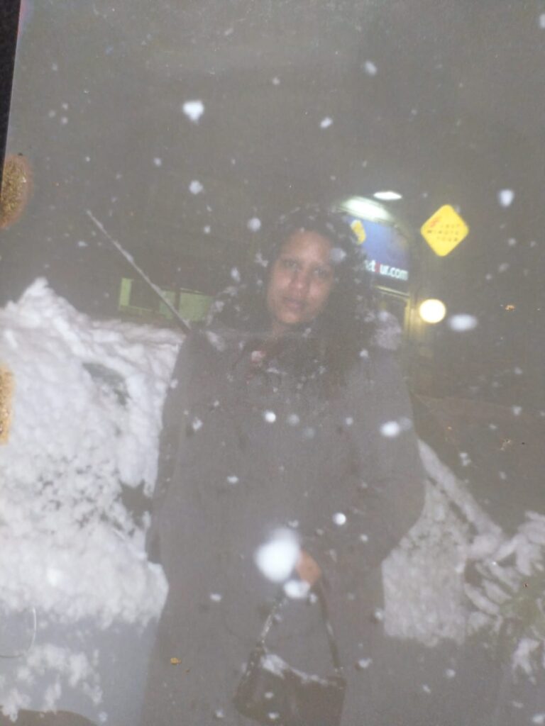 Foto analógica digitalizada. A foto está com algumas manchas no canto esquerdo. No centro da foto, Kelly Passos com casaco corta vento e bolsa preta da cintura para cima. É possível ver muita neve ao redor dela e atrás dela, bem como na frente da lente da câmera.