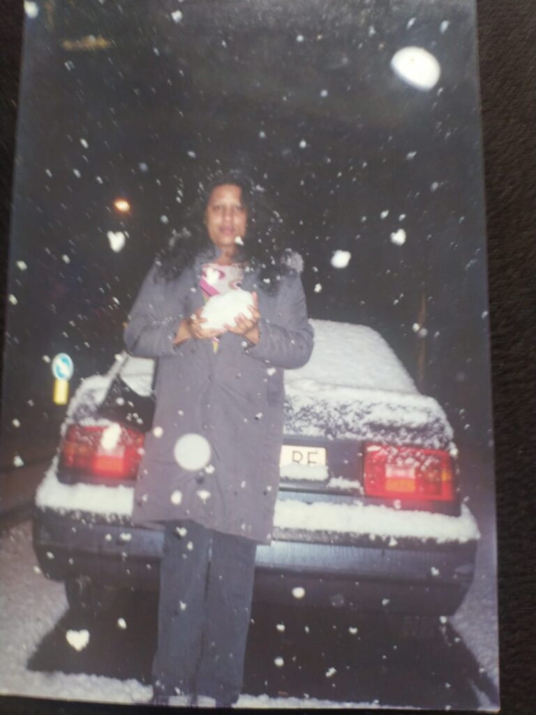 Foto analógica digitalizada, bastante deslocada do quadro. É possível ver grandes porções do tampo da mesa na digitalização. No centro da foto Kelly Passos veste casaco corta-vento, segurando uma bola de neve com as mãos no centro do peito. Ela está na frente de um carro. É possível ver neve sobre o carro, no chão, e caindo ao redor da pessoa e na frente da lente da câmera.